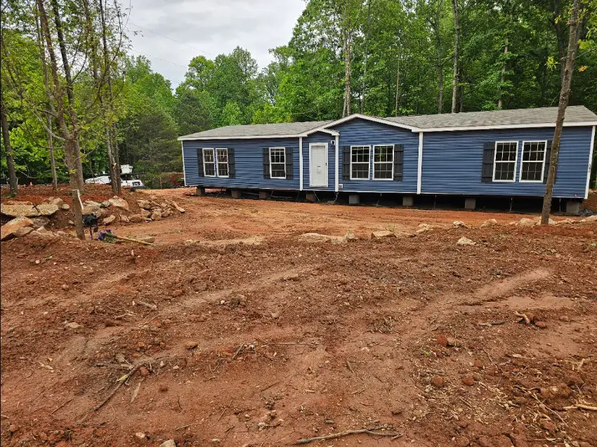 Blue mobile home on piers with rough grading and rocks in the foreground